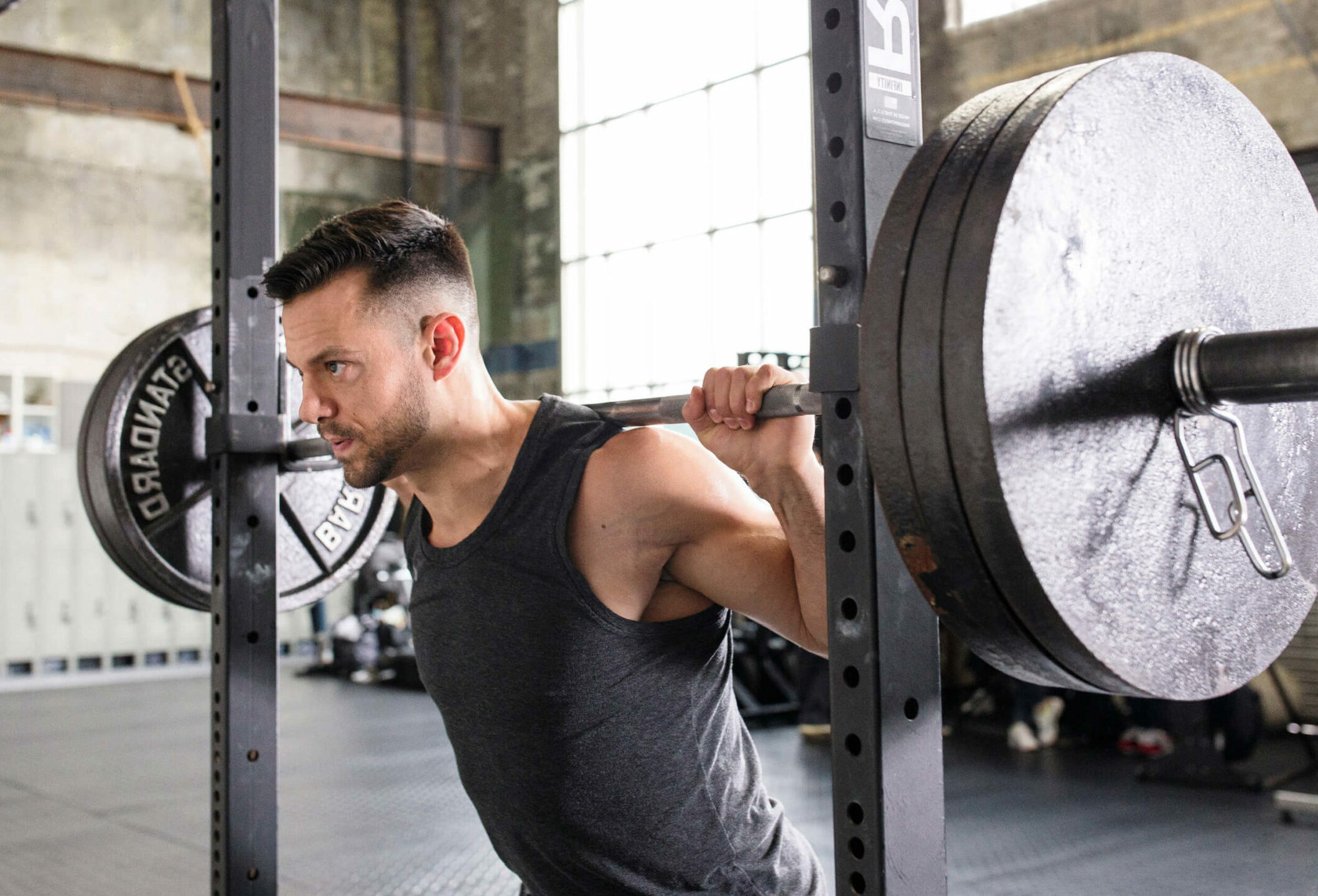 A picture of a Caliber online personal training client. A muscular male is wearing a grey vest performing a barbell back squat in a squat rack.